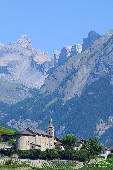 St-Séverin Church among green vineyards in Conthey, with steep rocky Alpine ridges behind it under a clear blue sky.