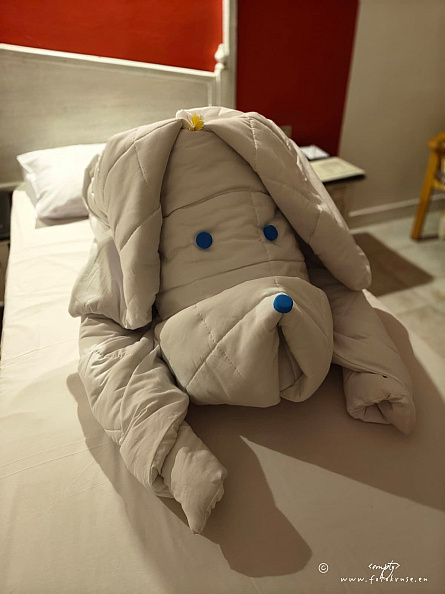 Folded white towel dog arranged on a hotel bed with blue bottle-cap eyes and nose and a small flower.
