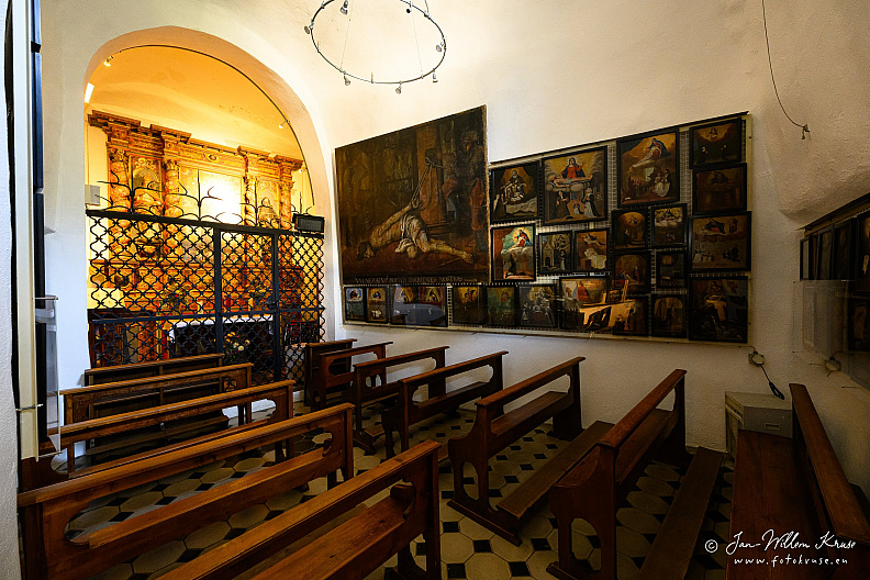 Small chapel interior with wooden benches, a metal grille and walls densely covered with painted ex-votos.