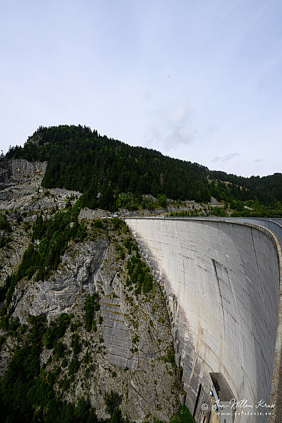 Tall curved concrete arch dam viewed obliquely beside steep grey rock and forest.