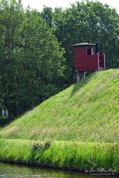 Small red wooden elevated outhouse structure on a grassy fortress rampart above the moat.