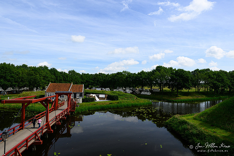 Red wooden drawbridge over a dark moat leading toward grassy ramparts and white fort buildings.