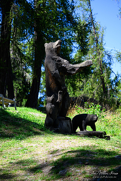 Large dark wooden carving of an upright bear with smaller bear figures beside a forest path.