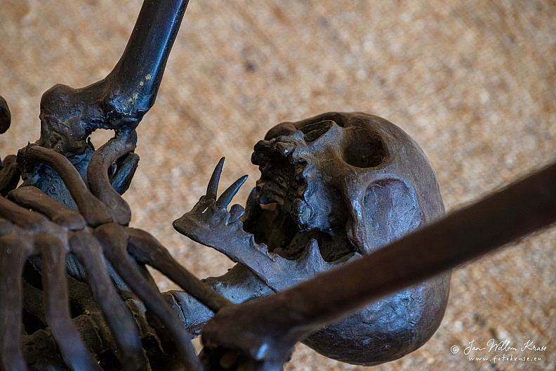 Close opposite-side view of the skull and jaw of André Lassen's bronze Spider sculpture, framed by skeletal limbs and rib bones.