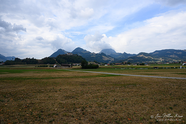 Wide landscape of the Plaine des Marches with Château de Gruyères small on its hill and pre-Alpine mountains behind.